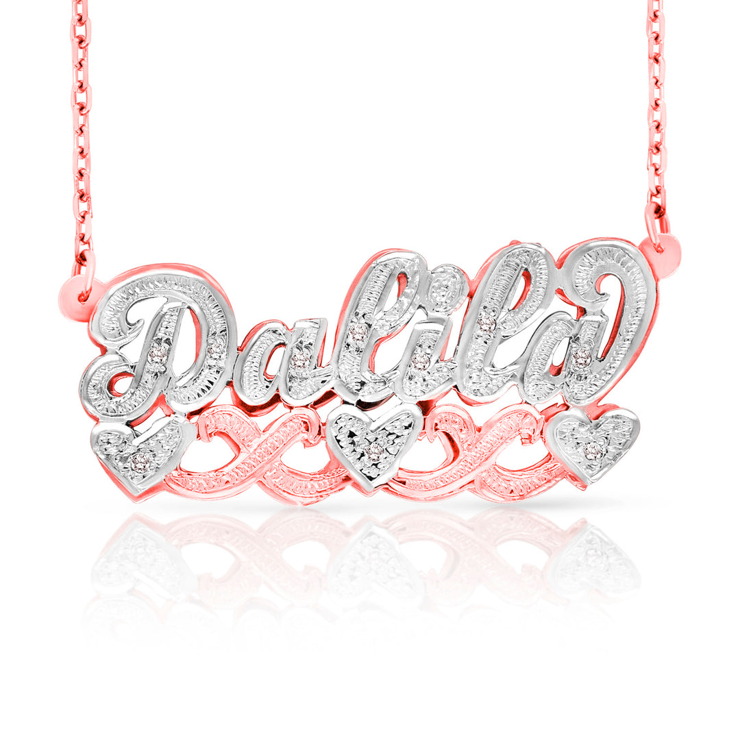 A personalized name necklace reading 'Dalia' in silver-tone script with clear stones, set over a rose gold chain and matching heart-shaped accents, shown on a white background with a reflection.