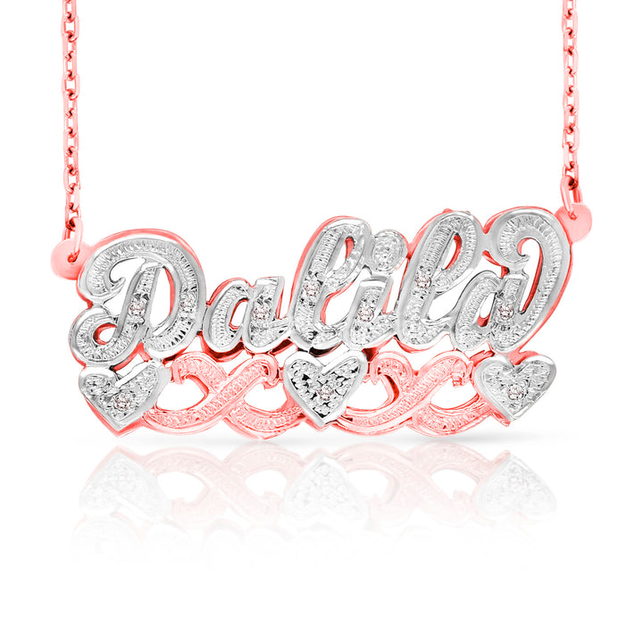 A personalized name necklace reading 'Dalia' in silver-tone script with clear stones, set over a rose gold chain and matching heart-shaped accents, shown on a white background with a reflection.